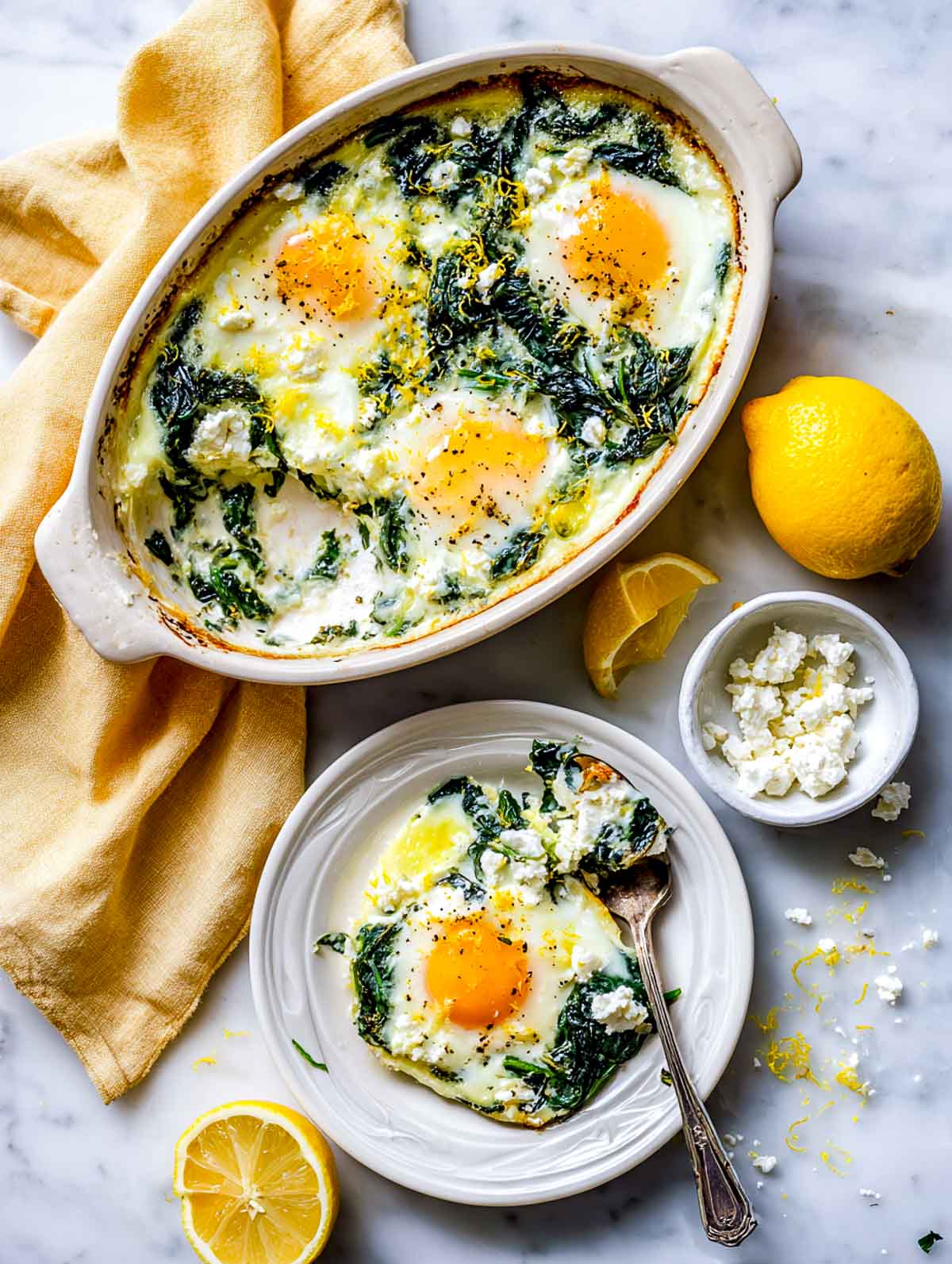 Top-down view of baked eggs with feta and spinach in an oval ceramic baking dish on a white marble surface, with one portion scooped out and served on a white plate, showing softly set eggs, wilted spinach, crumbled feta, and lemon zest, with lemon halves and a yellow napkin nearby.