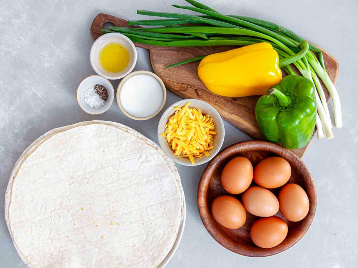 Ingredients for breakfast burritos including eggs, tortillas, peppers, cheese, scallions.