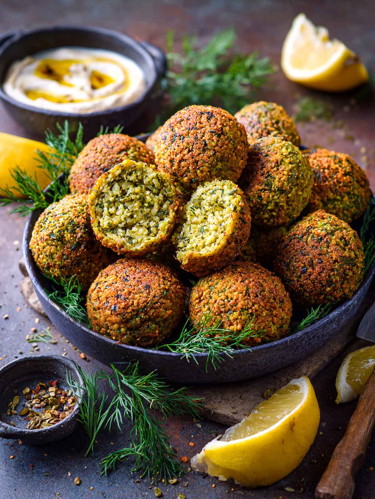 Crispy baked fava bean falafel with fresh herbs and lemon wedges placed in a black bowl with one broken falafel.