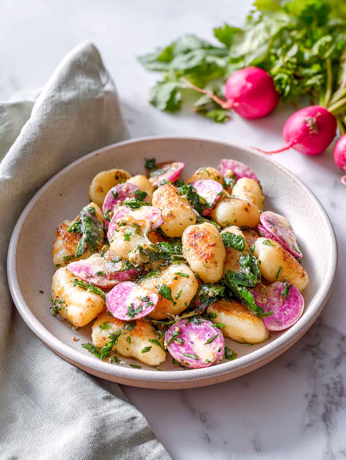 A shallow bowl of crispy pan-roasted gnocchi tossed with caramelized radish slices and fresh herbs, lightly coated in a warm herb vinaigrette on a white marble surface.