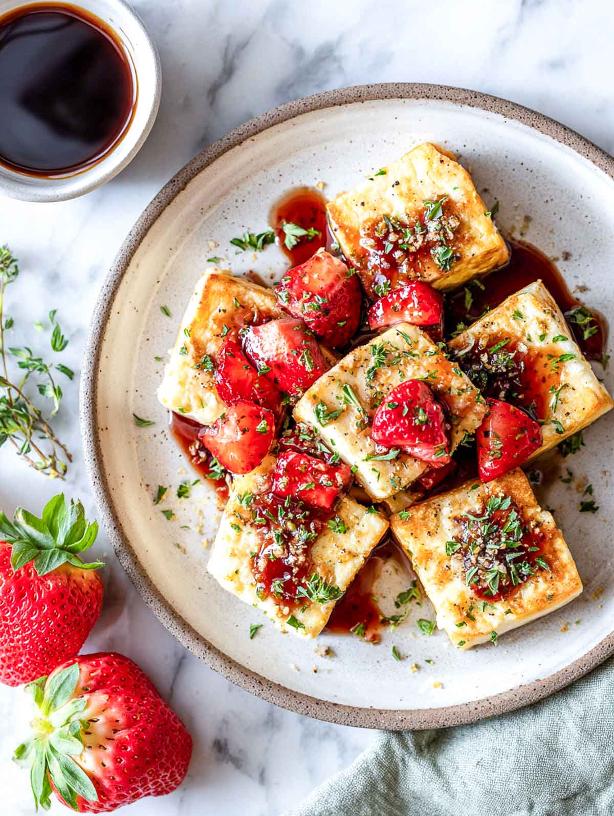 Crispy pan-seared tofu cubes topped with a glossy strawberry balsamic sauce and fresh herbs, served on a ceramic plate with whole strawberries nearby.