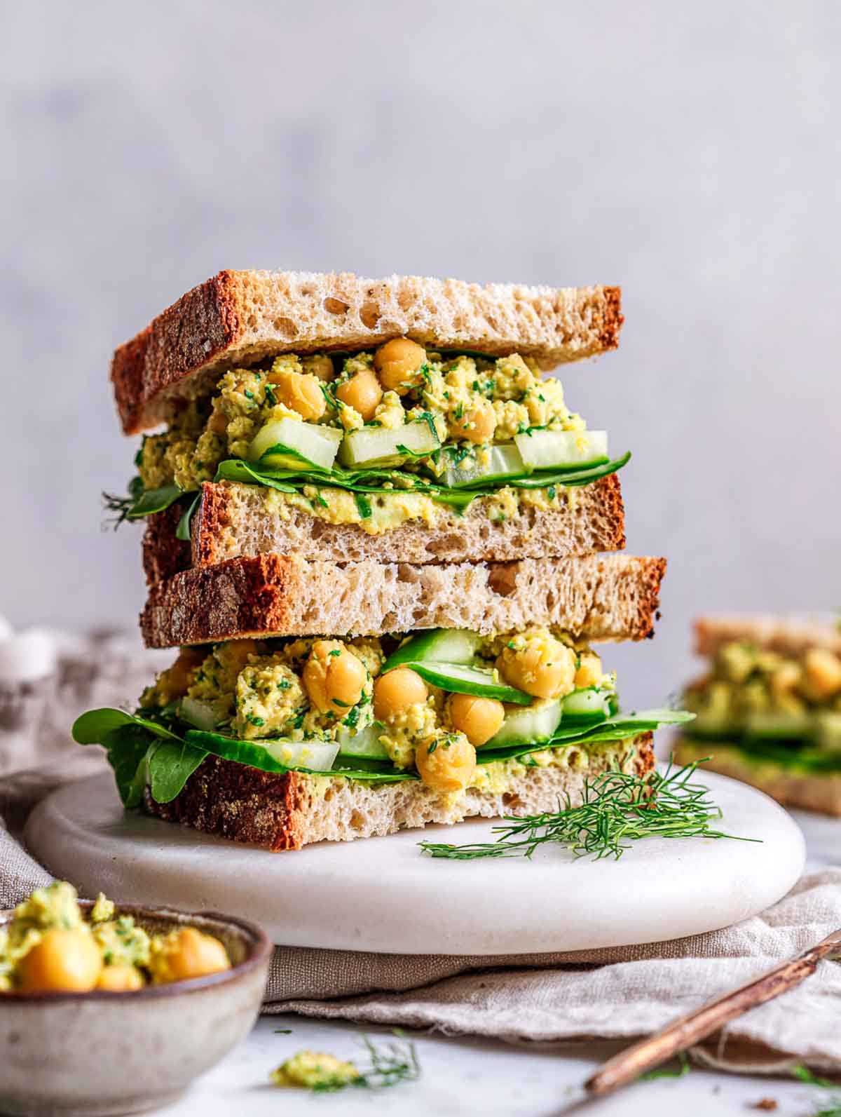 Stacked sandwich filled with chunky chickpea smash, fresh herbs, cucumber slices, and leafy greens on rustic bread.