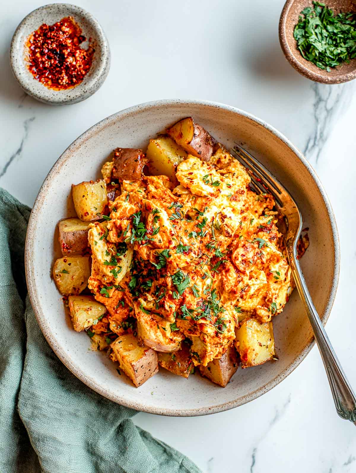 Top-down view of harissa scrambled eggs served with roasted potato chunks in a wide ceramic bowl on a white marble surface, showing soft scrambled eggs coated in red harissa, golden potatoes, chopped fresh herbs on top, and a small bowl of chili paste nearby.