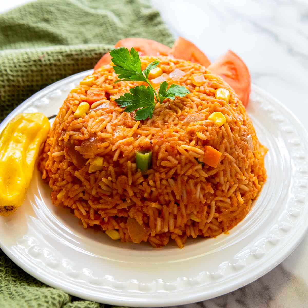 Close-up of jollof rice molded on a white plate, showing fluffy tomato-seasoned rice with vegetables, garnished with parsley and served with tomato slices and a whole pepper.