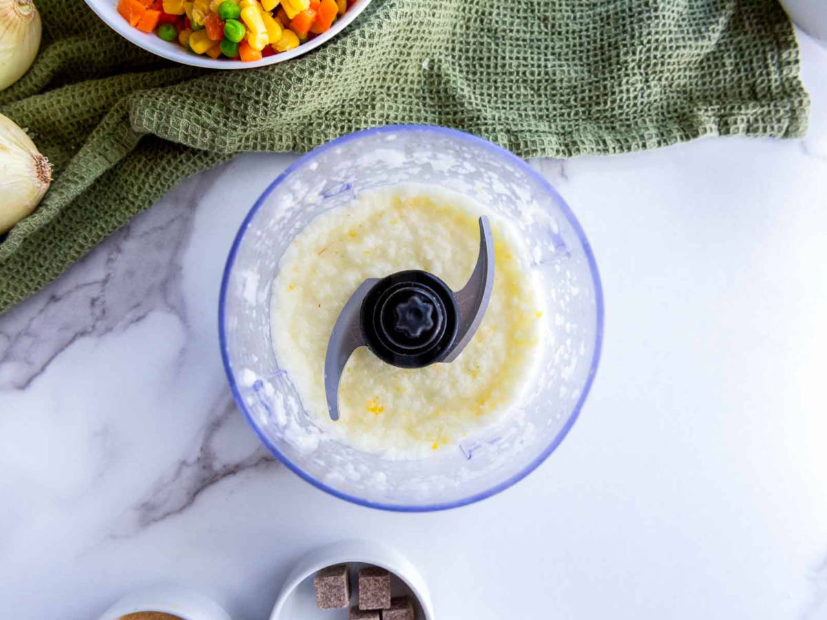 Overhead view of blended onion mixture in a food processor bowl on a marble surface, with jollof rice ingredients partially visible nearby.