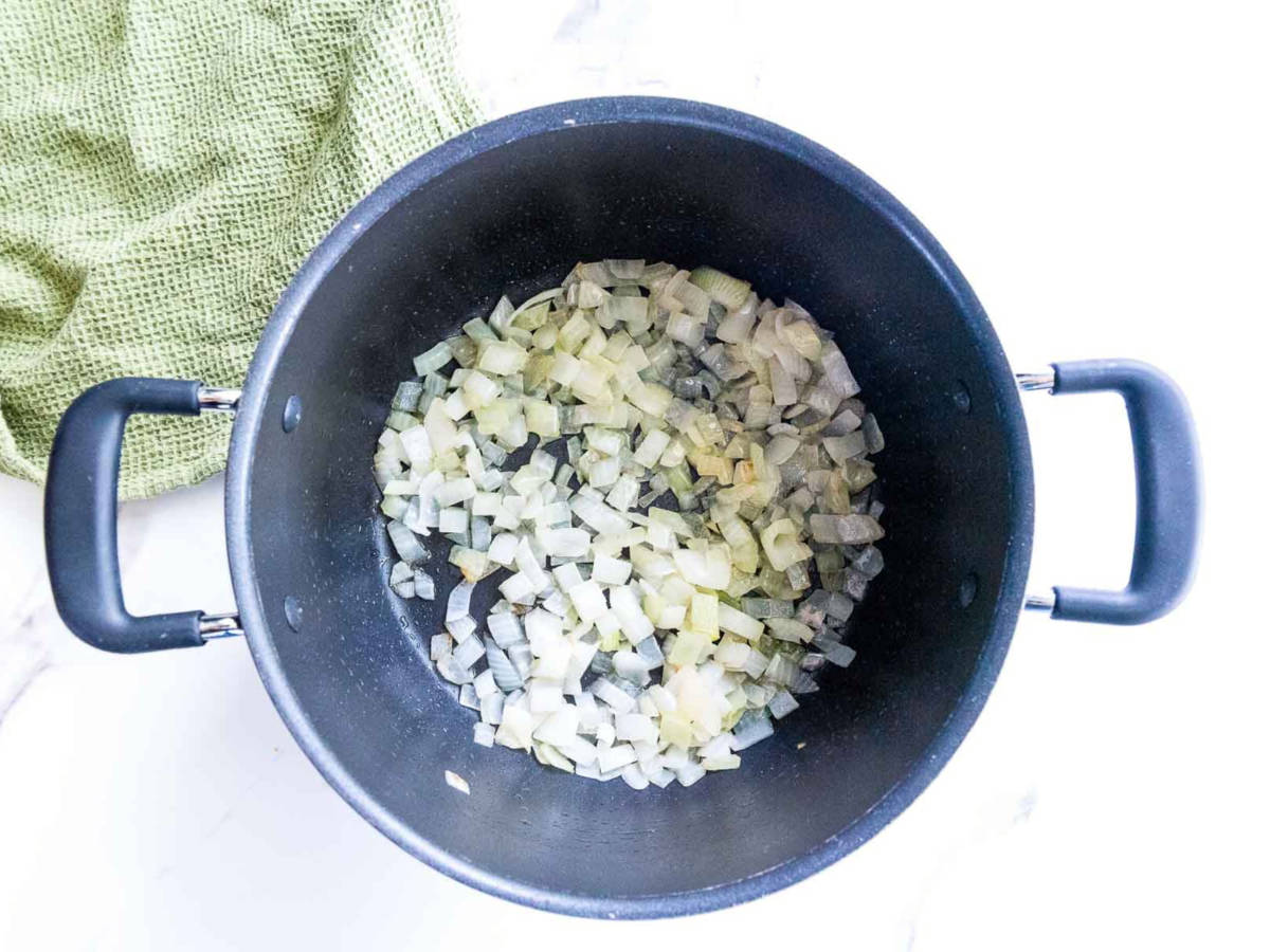 Overhead view of diced onions sautéing in a large pot, beginning to soften and turn translucent on a stovetop.