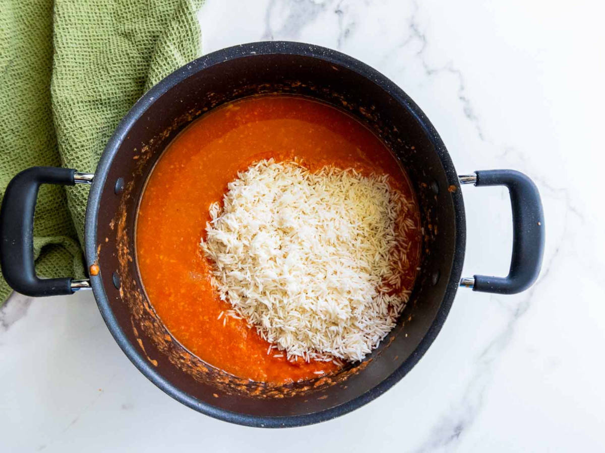 Uncooked white rice added to the pot of tomato sauce, ready to be stirred and cooked for jollof rice.