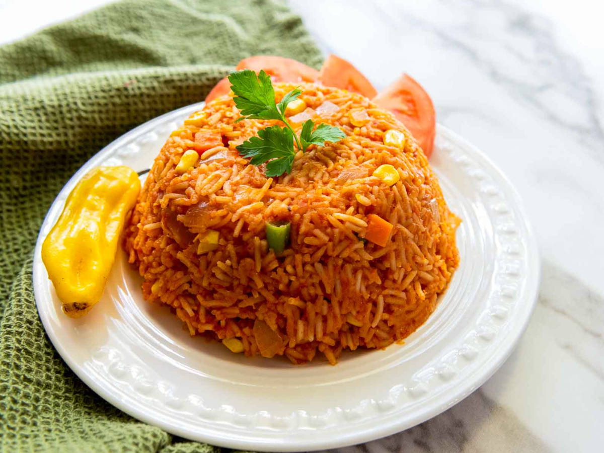 Plated jollof rice molded into a dome, garnished with parsley and served with sliced tomatoes and a whole pepper on the side.