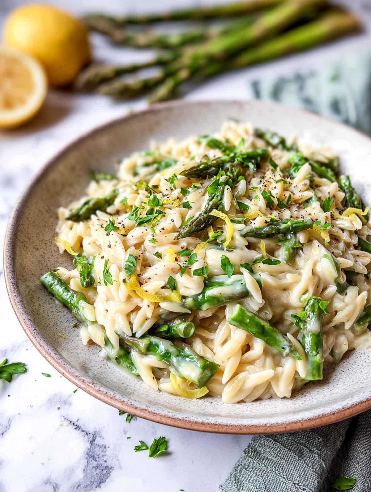 A shallow bowl of lemon asparagus orzo coated in a creamy white bean sauce, finished with fresh parsley, cracked black pepper, and lemon zest on a white marble surface.