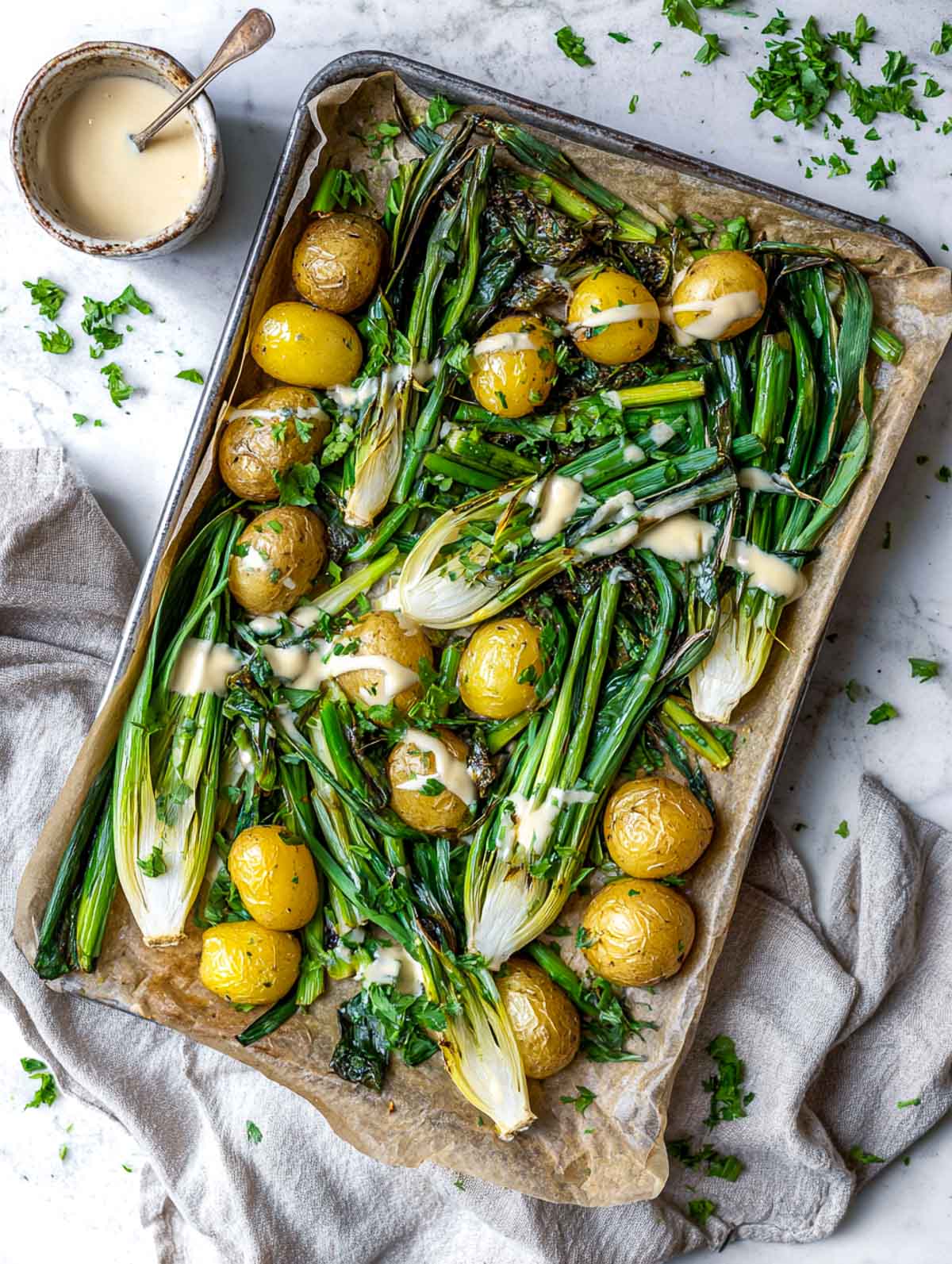 A sheet pan of roasted spring onions with long green tops and golden baby potatoes drizzled with creamy lemon tahini sauce.