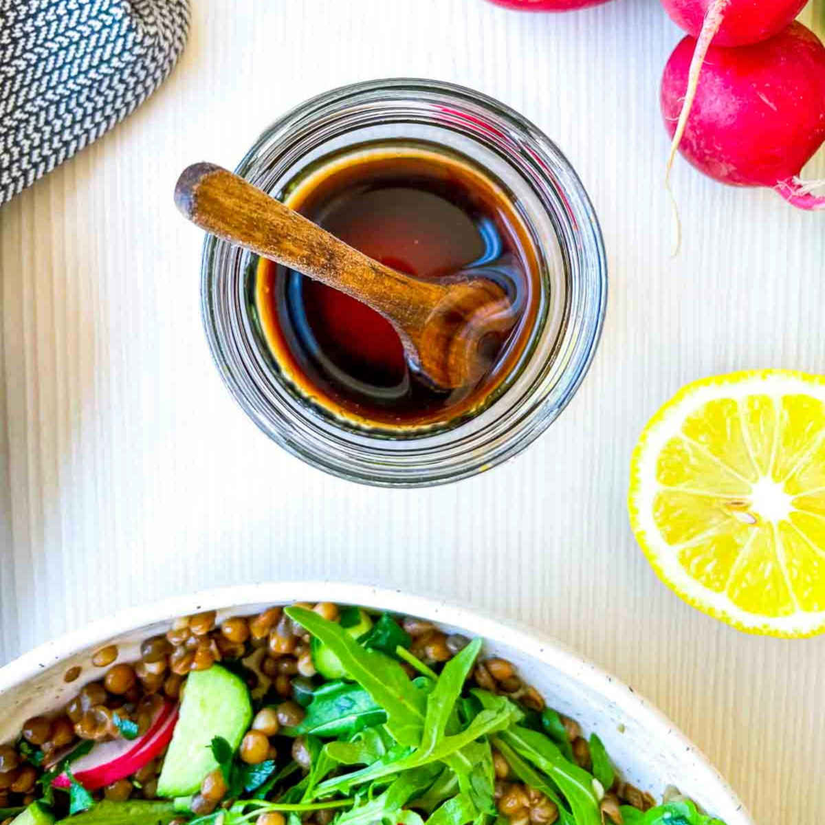 Overhead view of a small glass jar filled with dark vinaigrette and a wooden spoon, placed above a bowl of lentil salad with arugula, cucumber, and radishes, with lemon and radishes nearby.