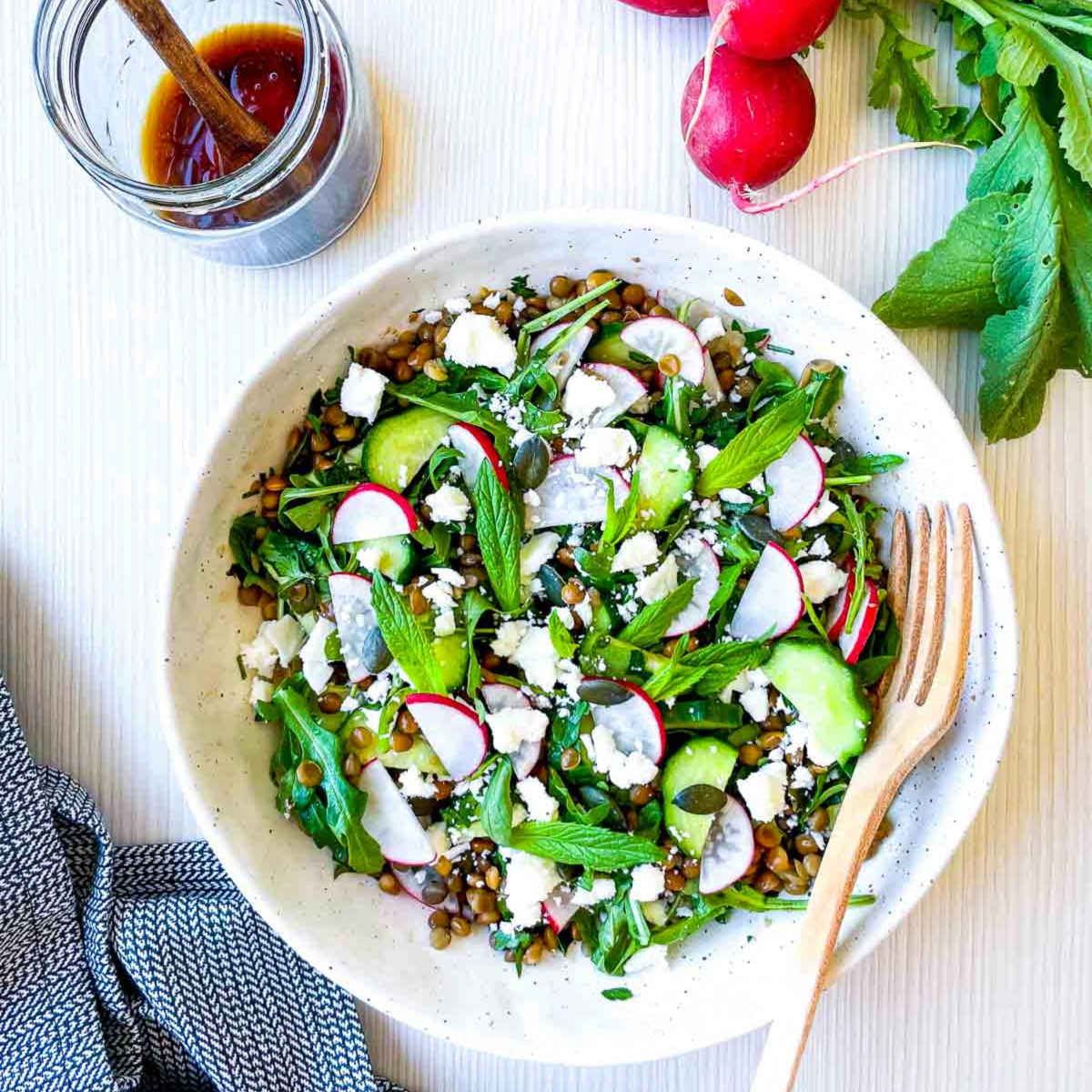 Overhead view of a lentil salad in a white bowl with arugula, cucumber slices, radishes, fresh herbs, crumbled feta, and seeds, with a jar of dressing and wooden fork on the side.