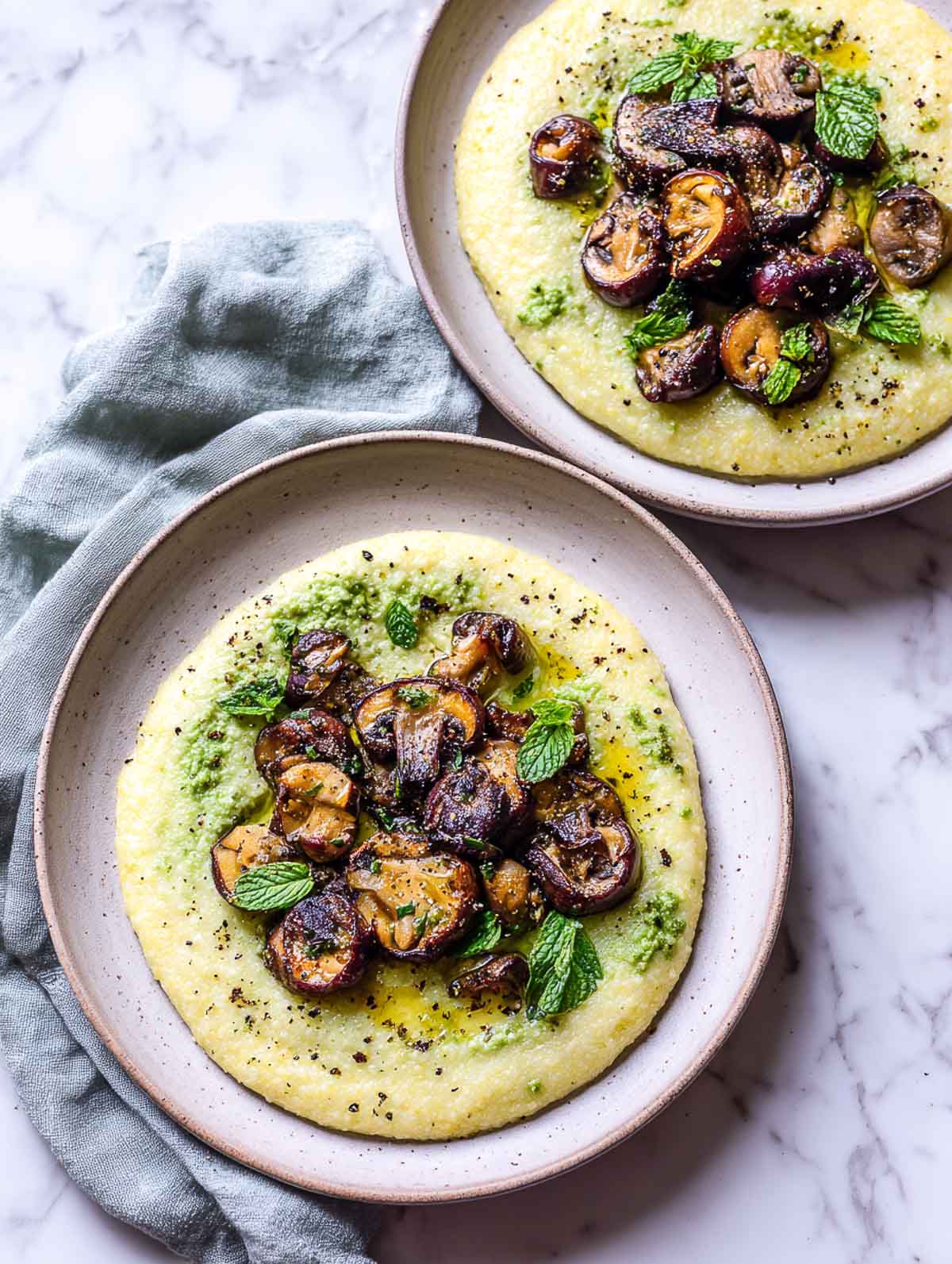 Two plates of creamy pea and mint polenta topped with deeply roasted mushrooms, fresh mint leaves, and cracked black pepper on a white marble surface.