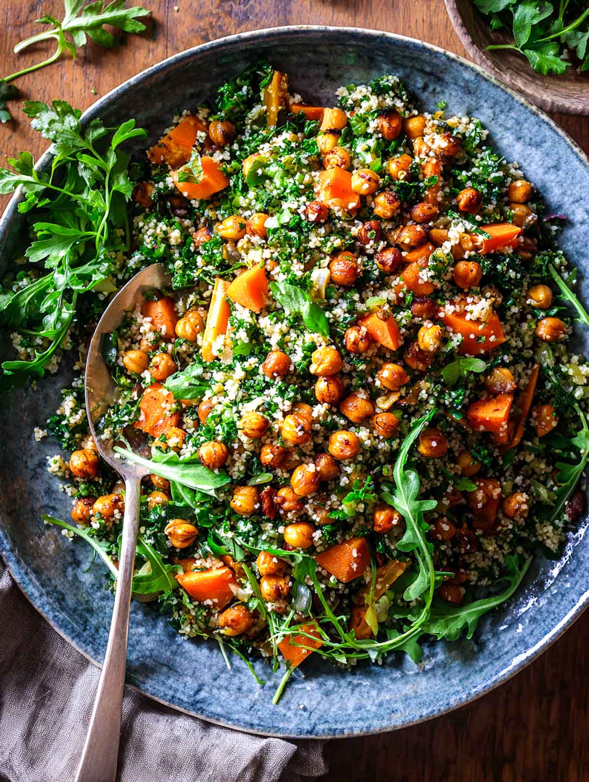 A bowl of roasted carrot and chickpea tabbouleh with finely chopped parsley, lemon dressing, and fluffy bulgur, shown with a serving spoon on a dark ceramic plate.