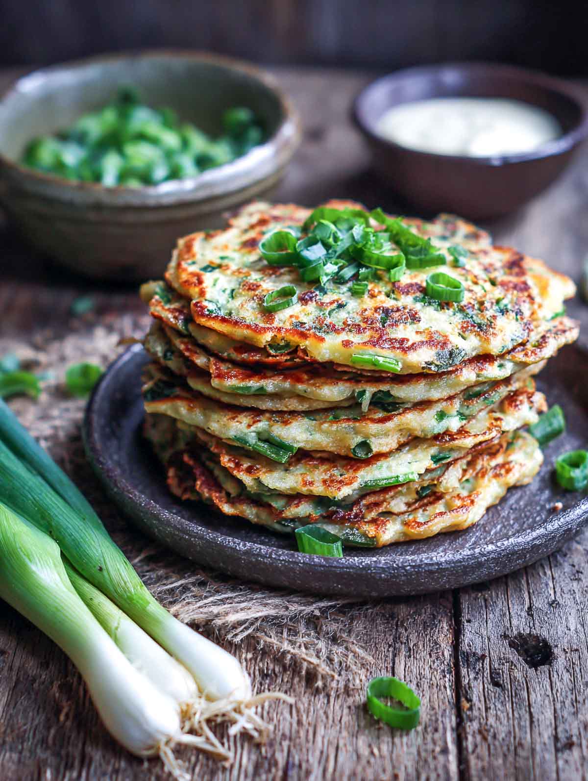 A stack of golden savory scallion pancakes made with oat flour, topped with sliced green onions and served on a rustic plate with dipping sauce.