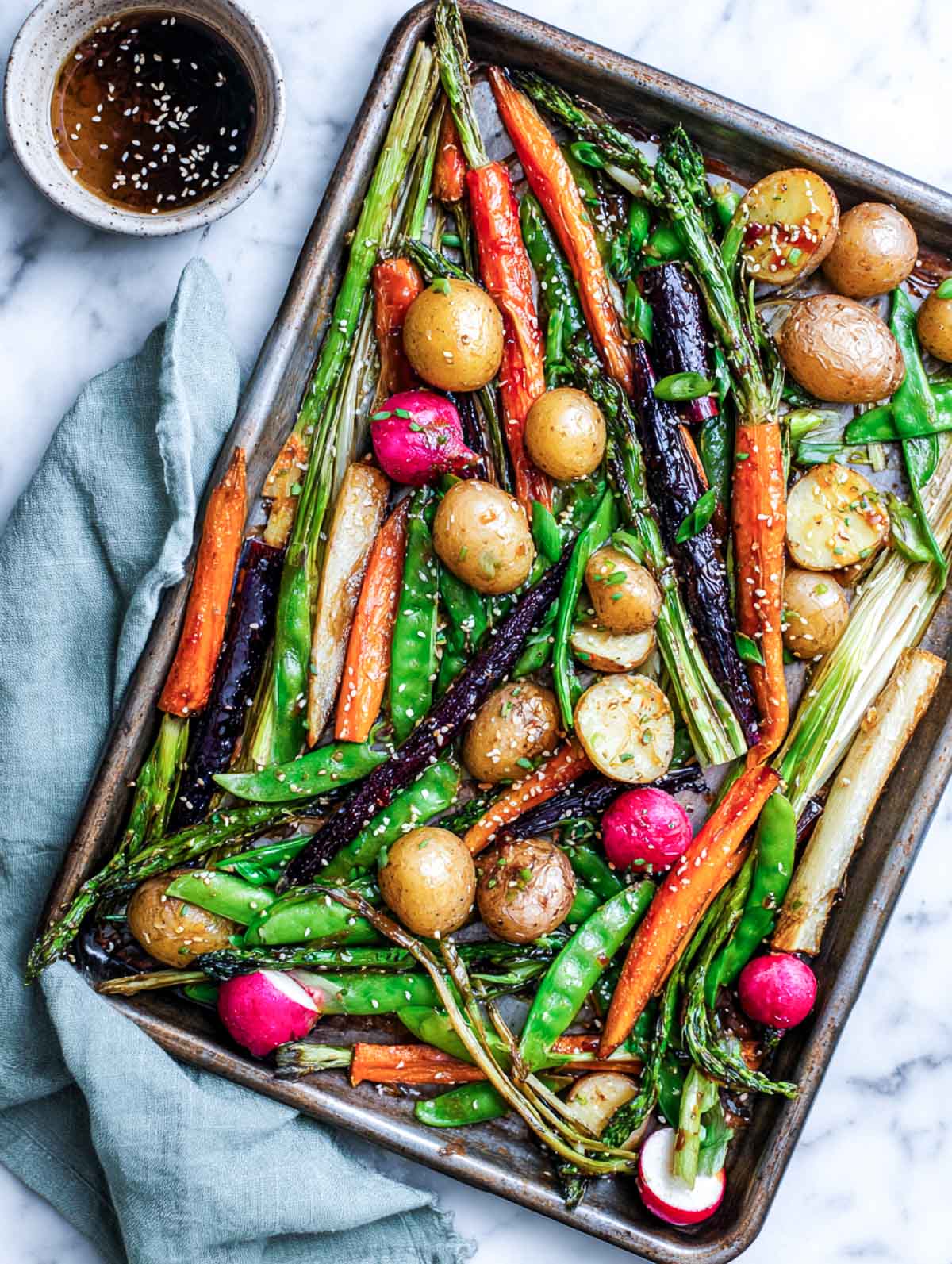 A top-down view of a sheet pan filled with roasted spring vegetables glazed with miso maple sauce, including asparagus, carrots, snap peas, baby potatoes, and radishes on a white marble surface.