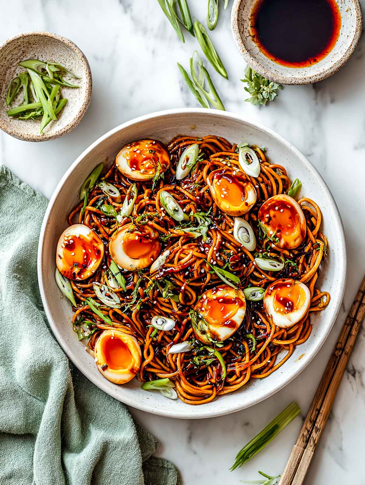 Top-down view of soy glazed jammy eggs served over scallion noodles in a wide ceramic bowl on a white marble surface, with halved eggs showing soft jammy yolks, glossy noodles coated in soy sauce, sliced scallions scattered on top, and a small bowl of extra soy glaze nearby.