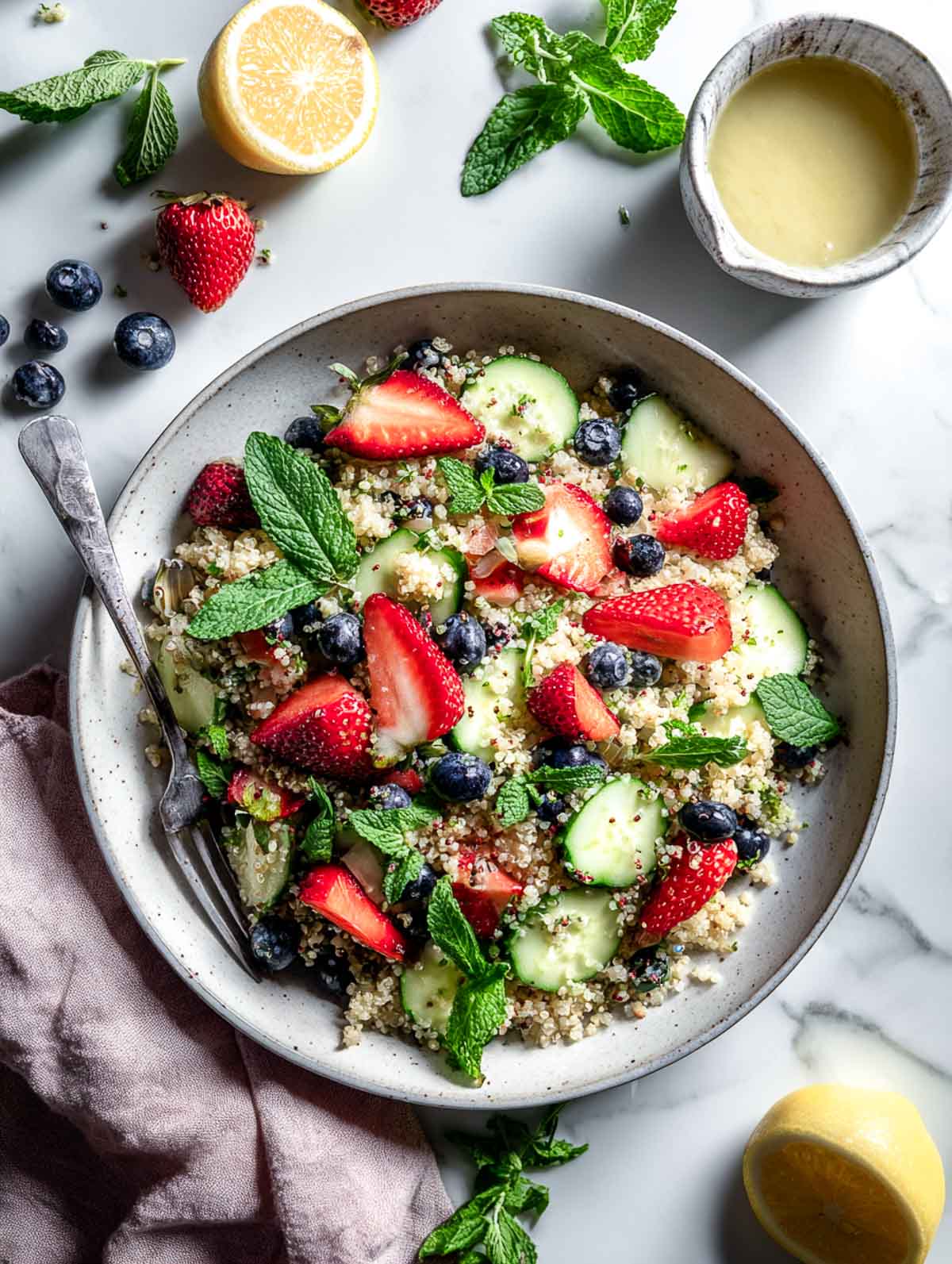A bowl of spring quinoa salad with sliced strawberries, blueberries, cucumber, fresh mint, and lemon dressing on a marble surface.