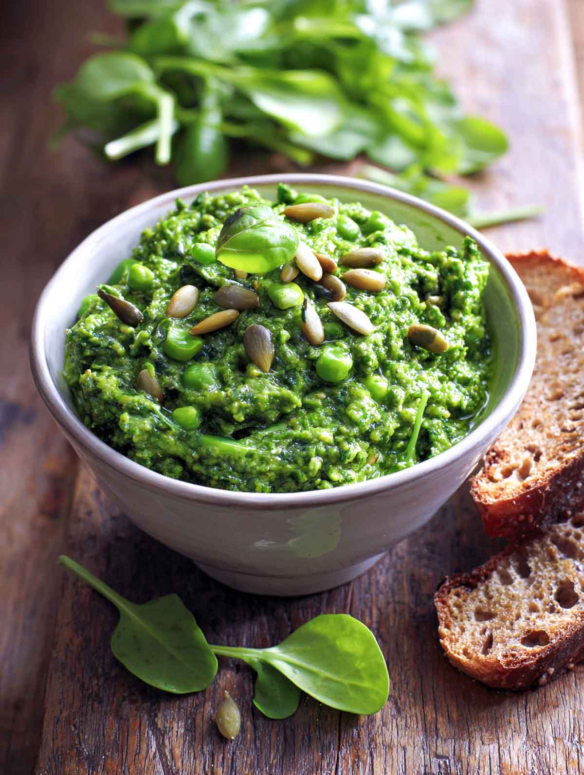 A bowl of vibrant green spring pesto made with spinach, peas, and sunflower seeds, garnished with fresh basil and served with slices of rustic bread on a wooden surface.