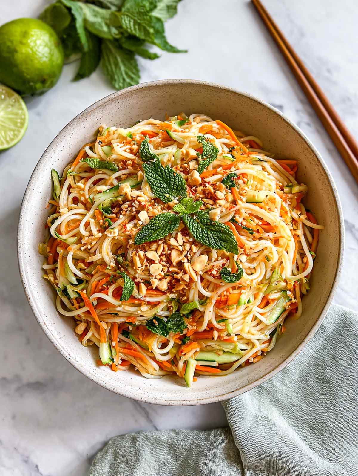 A top-down view of a bowl of spring roll salad made with rice noodles, shredded vegetables, fresh herbs, and crushed peanuts, served with lime and chopsticks on a white marble surface.