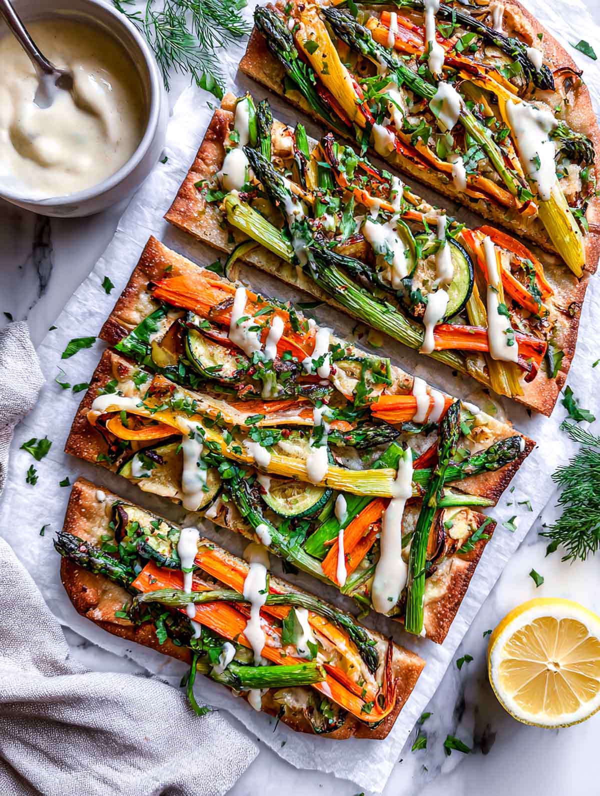Sliced rectangular flatbread topped with roasted spring vegetables and finished with a creamy lemon cashew drizzle on parchment paper.