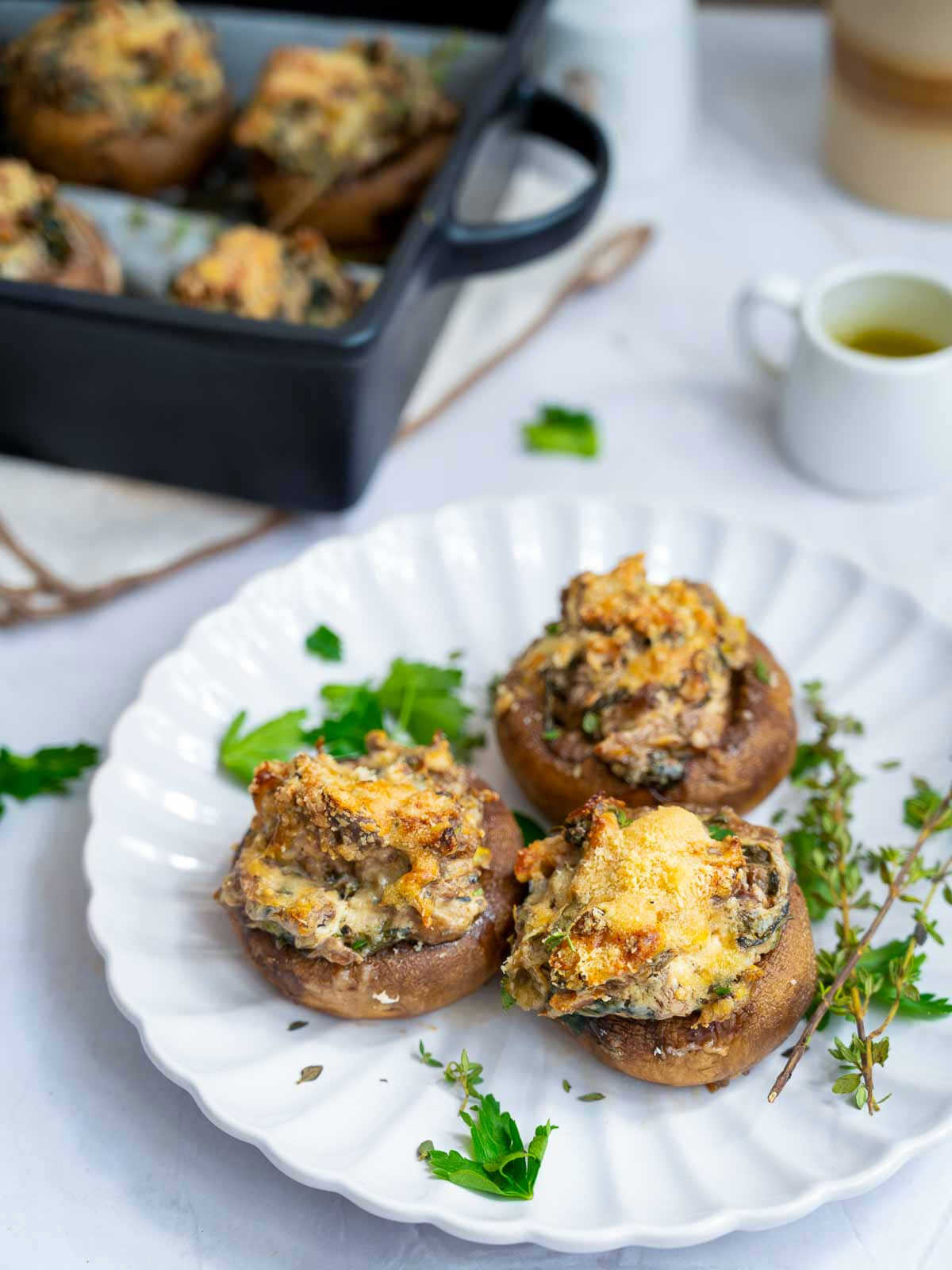 A plate of stuffed mushrooms placed on a whote marble surface with the baked dish in the background.