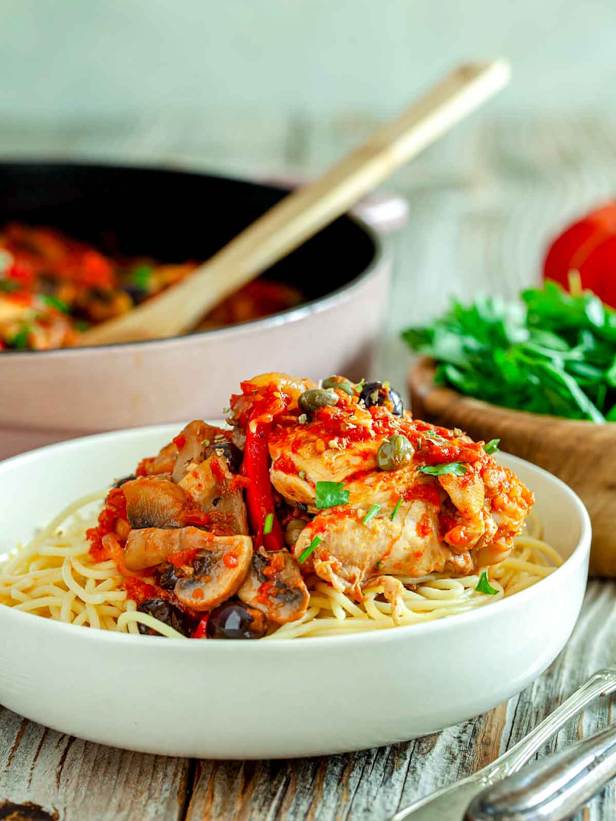A bowl of spaghetti topped with chicken cacciatore, mushrooms, olives, and bright tomato sauce, with the skillet in the background.