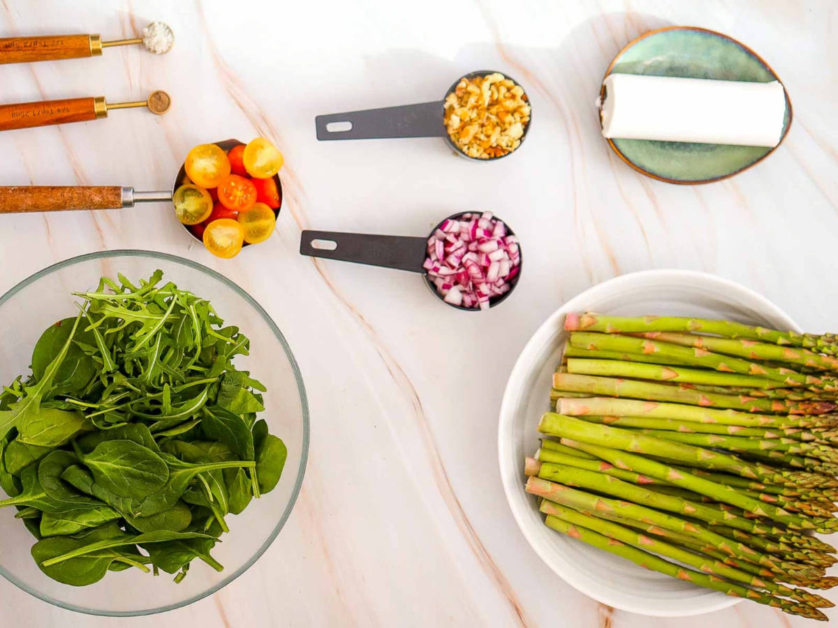 Ingredients laid out for asparagus goat cheese salad, including fresh asparagus, arugula and spinach, cherry tomatoes, chopped red onion, walnuts, and goat cheese