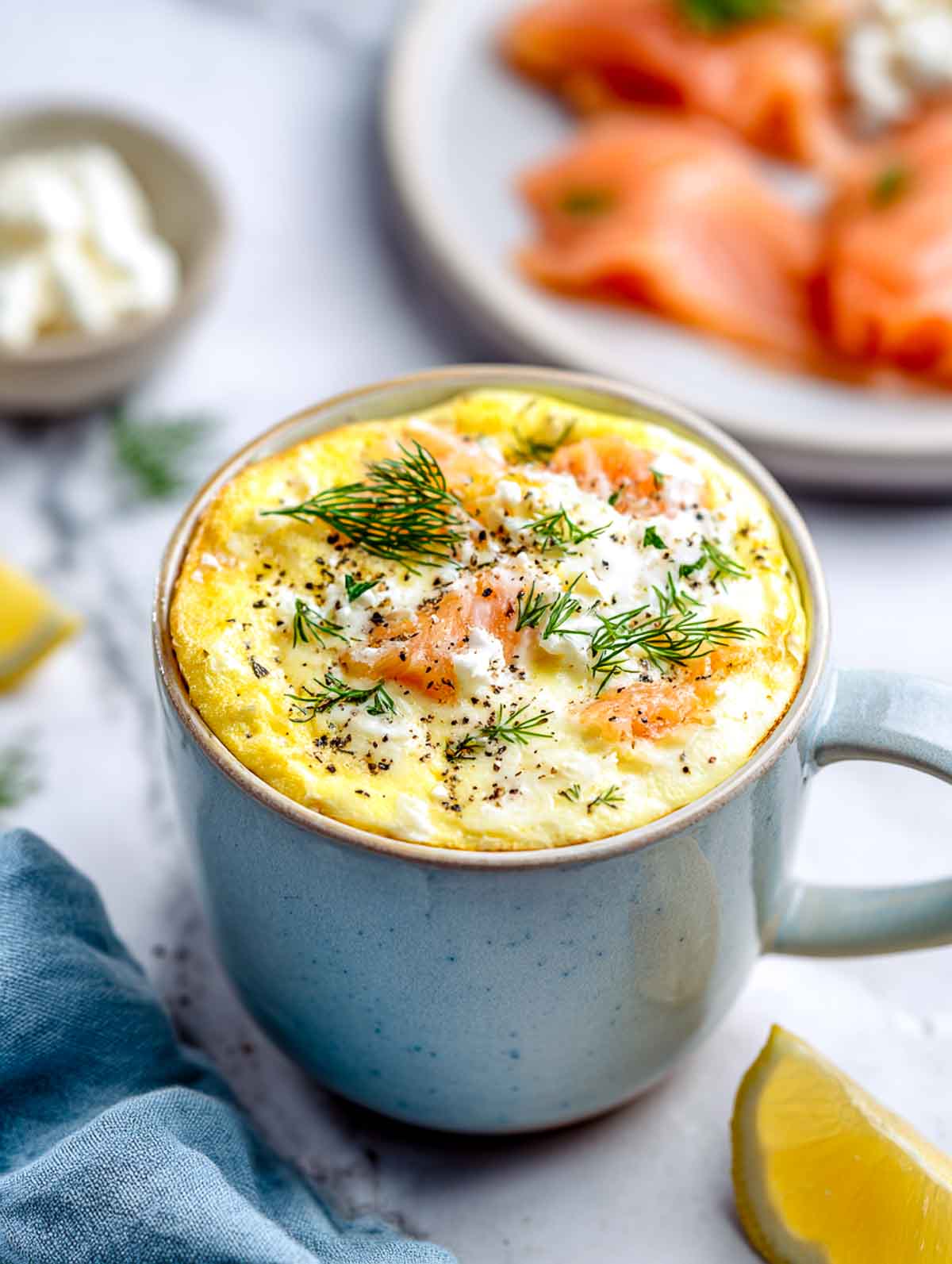 A light ceramic mug filled with a fluffy egg bake studded with smoked salmon and cottage cheese, topped with fresh dill and black pepper, set on a white marble countertop with lemon wedges and a blue napkin nearby.