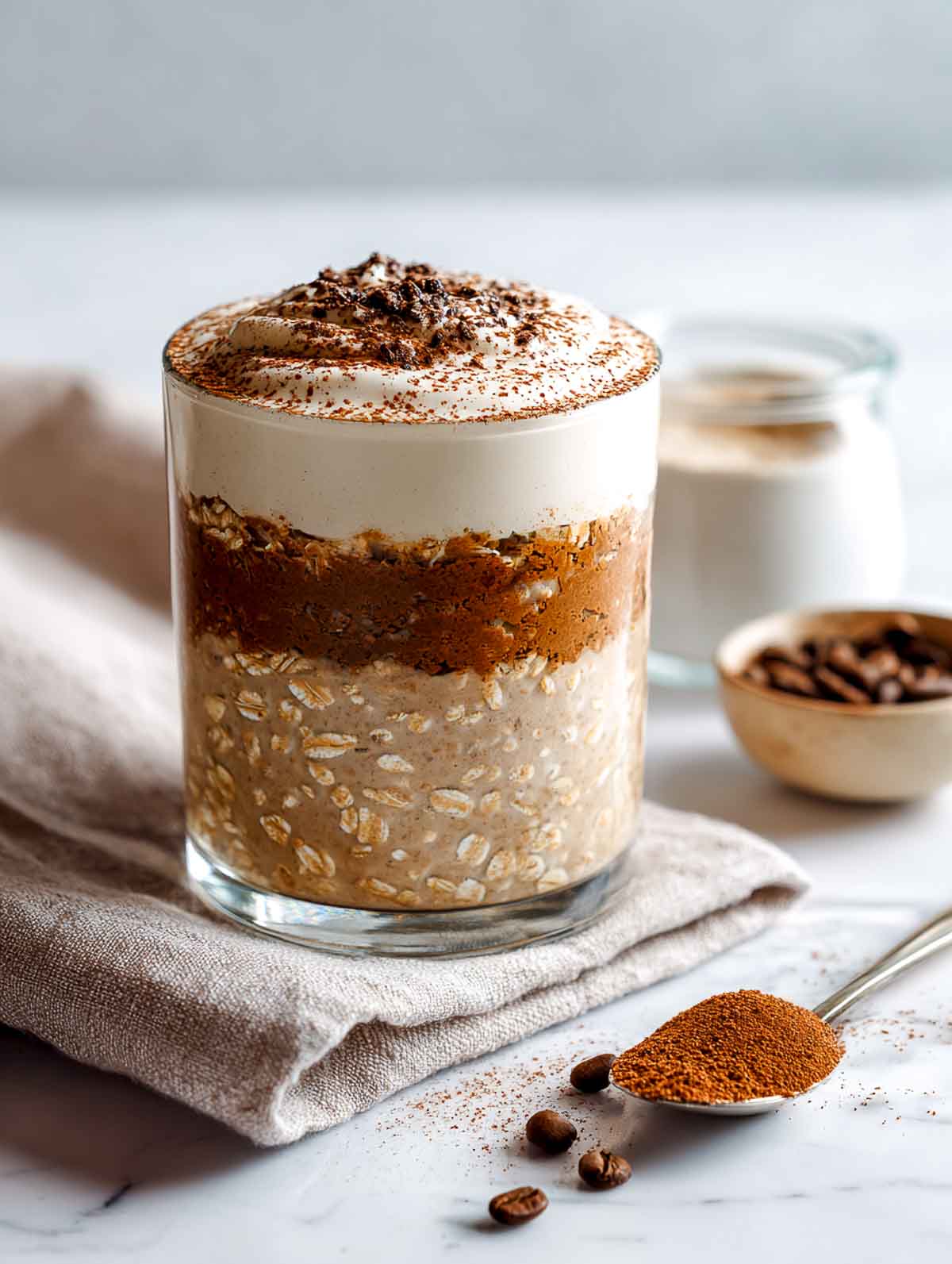 A bright angled view of a clear glass mug filled to the rim with layered tiramisu overnight protein oats, showing a light beige oat layer, a darker coffee-infused oat layer, and a thick creamy yogurt topping dusted with cocoa powder, set on a white marble countertop with a mocha linen napkin and espresso cup nearby.
