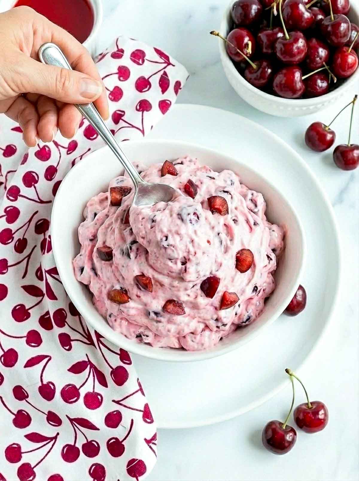 A spoon scooping cherry cheesecake fluff from a bowl, showing the light, airy texture with cherry pieces mixed throughout.