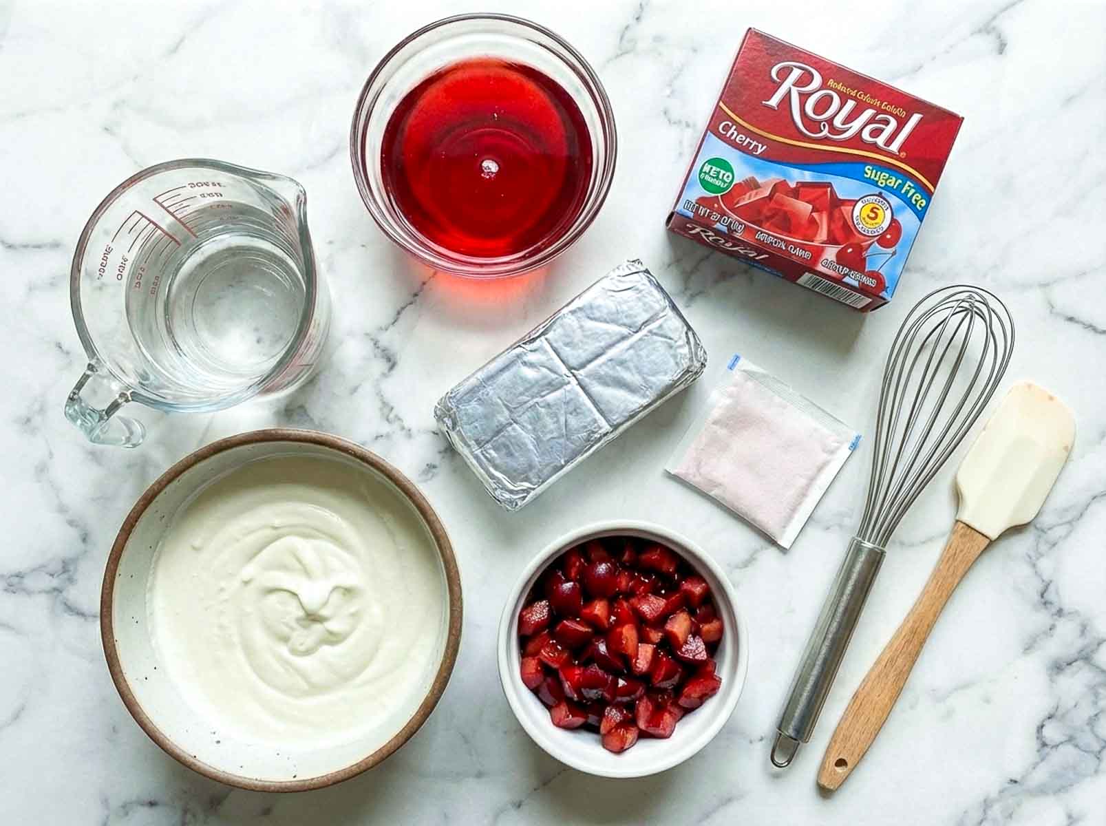 Ingredients for cherry cheesecake fluff including cream cheese, cherry gelatin, whipping cream, diced cherries, water, and mixing tools on a marble surface.