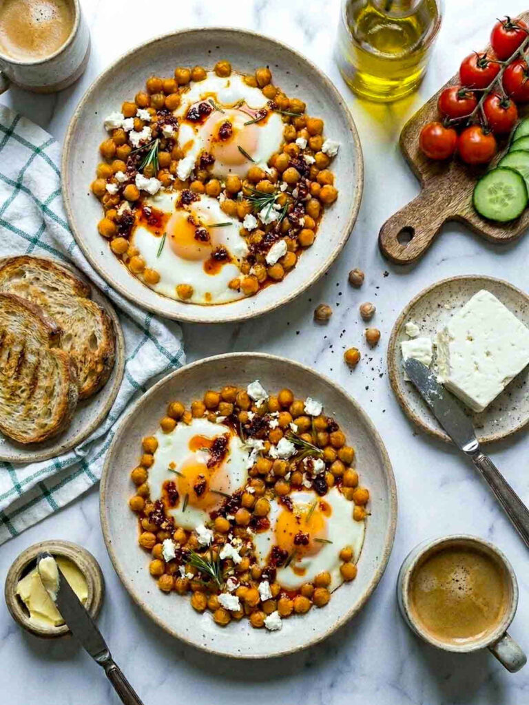 Overhead flat lay of two bowls of chickpea fried eggs with runny yolks, crumbled feta, chili oil, and fresh rosemary, served alongside toast, butter, coffee, cherry tomatoes, and sliced cucumber on a marble surface.