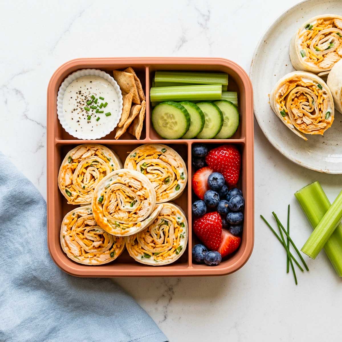 Buffalo ranch chicken pinwheels packed in a school lunchbox with celery, cucumber, berries, and ranch dip.