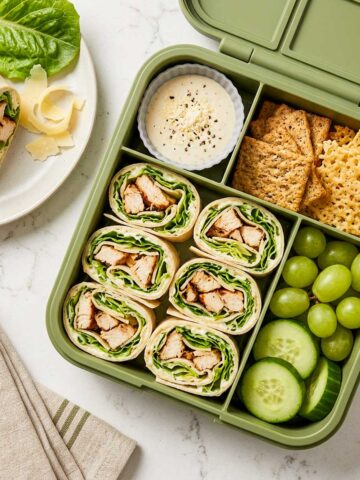 Chicken Caesar wrap slices packed in a school lunchbox with grapes, cucumber, crackers, Parmesan crisps, and Caesar dip.