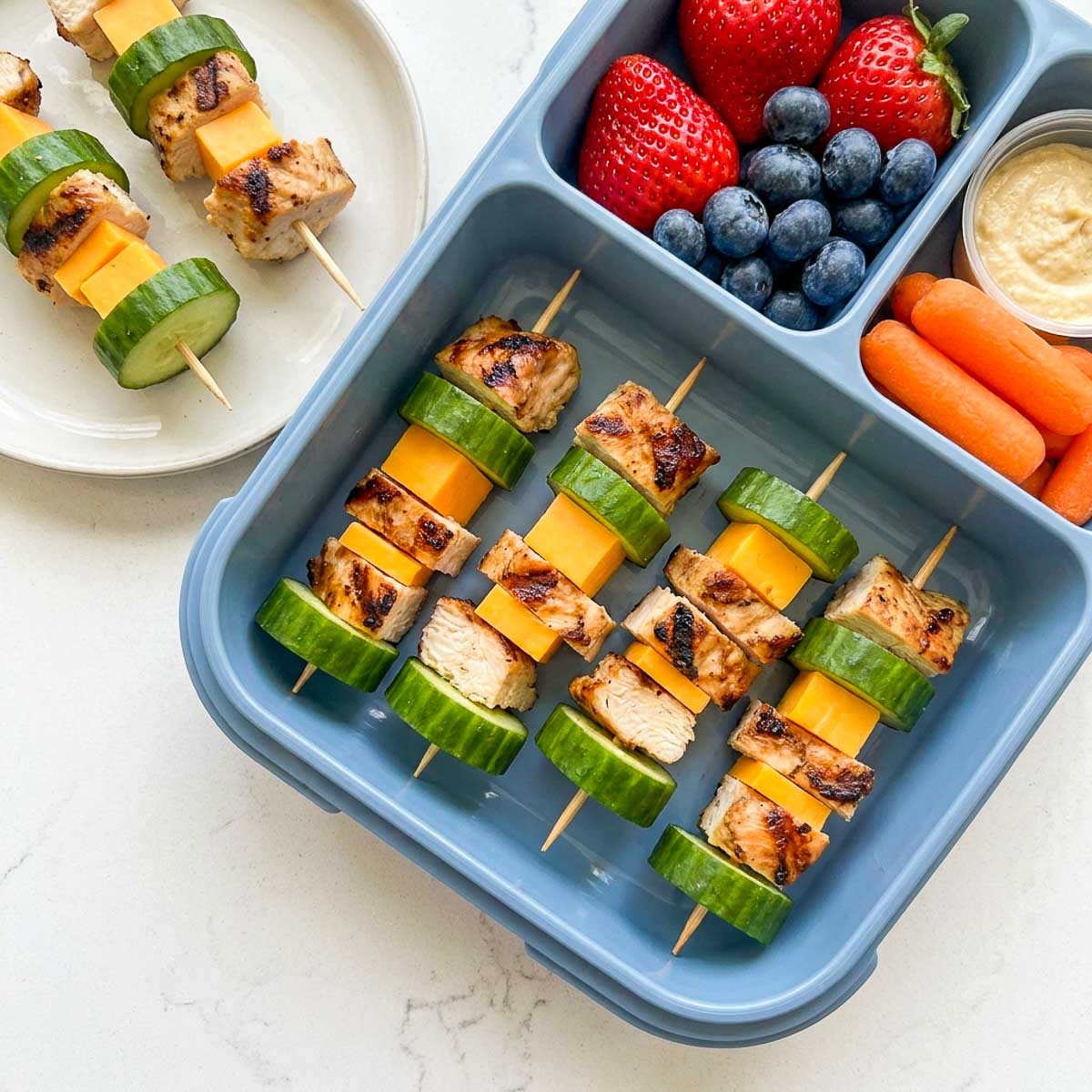Cold mini chicken skewers with cucumber and cheddar cheese packed with berries, baby carrots, and hummus.
