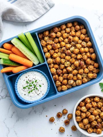 Crispy roasted chickpeas with ranch dip, cucumber, celery, and carrots in a blue lunchbox.