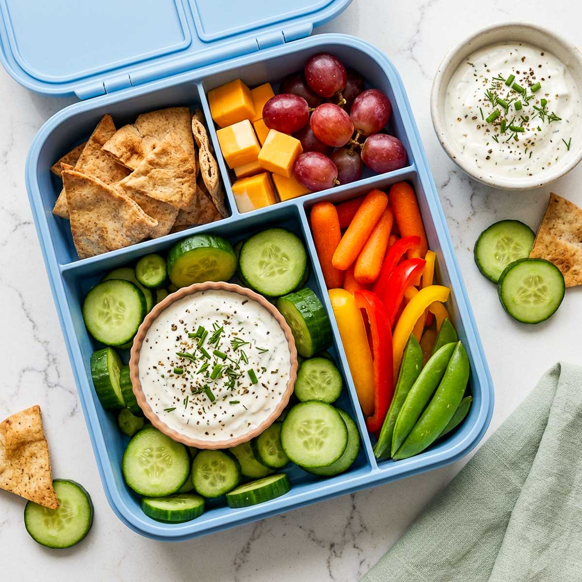 Greek yogurt ranch dip with cucumber coins packed with pita chips, cheese cubes, grapes, carrots, bell peppers, and snap peas.