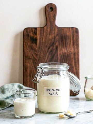 Homemade milk kefir in a labeled glass jar with a small glass of kefir and a jar of milk kefir grains beside it.