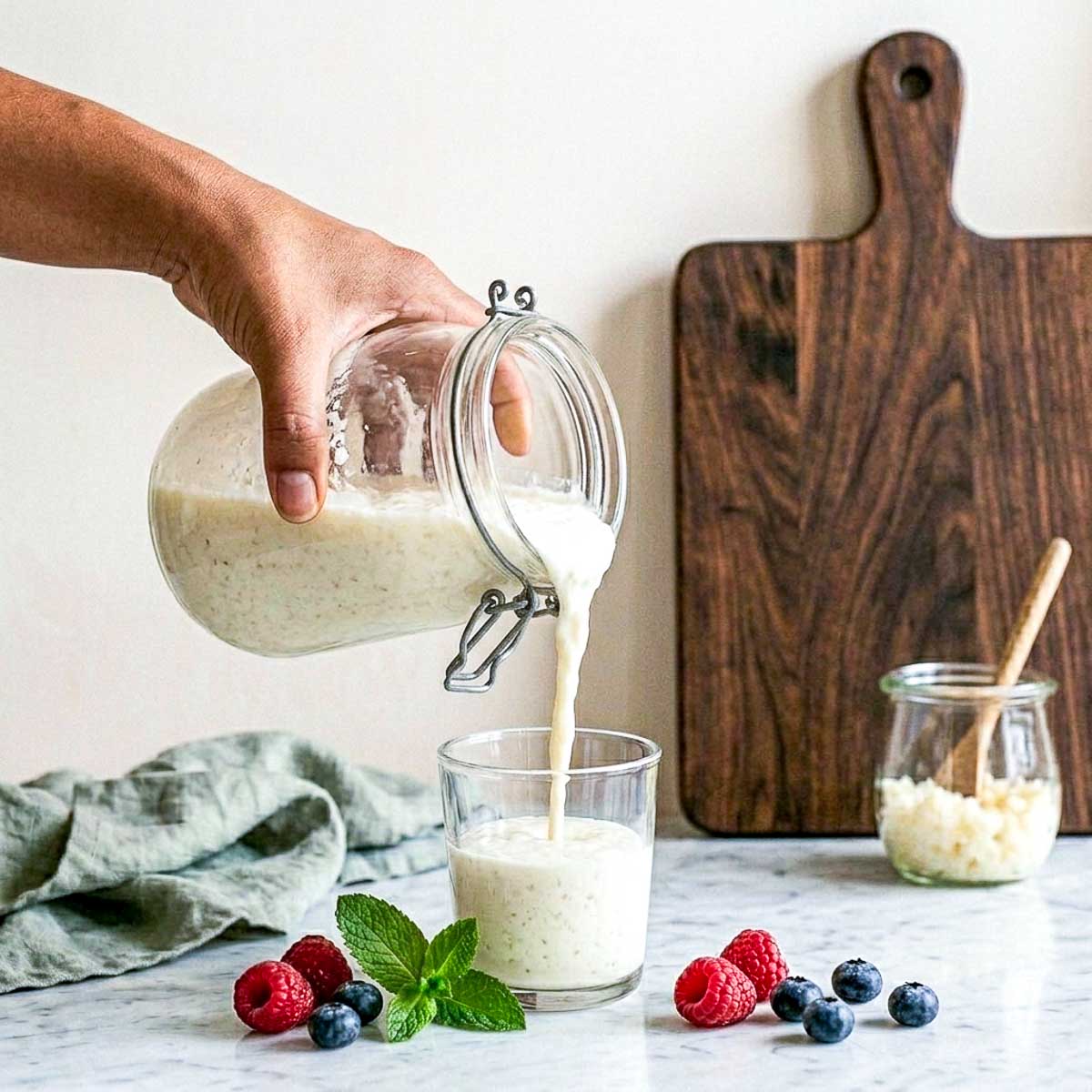 Pouring finished homemade milk kefir from a jar into a glass, with fresh berries and mint on the counter.
