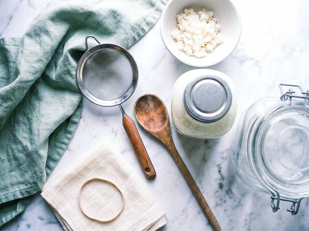 Everything needed to make milk kefir at home: kefir grains, milk, glass jar, fine mesh strainer, cloth cover, and rubber band.