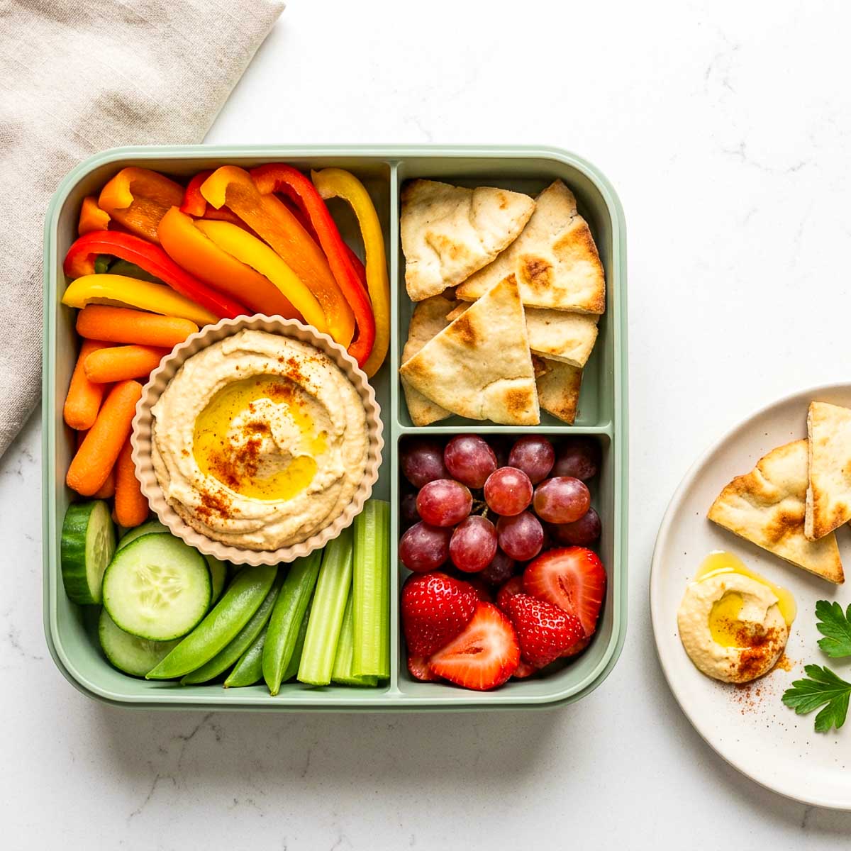 Hummus lunchbox with carrot sticks, cucumber, snap peas, bell peppers, pita chips, grapes, and strawberries.
