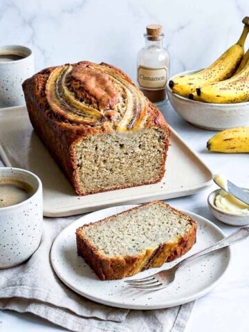 Sliced kefir banana bread loaf on a cream platter with a caramelized banana top, served with coffee and butter.