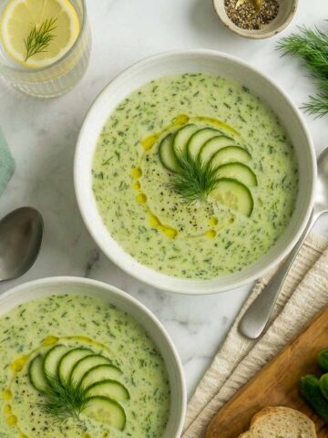 Two bowls of creamy kefir cucumber avocado soup garnished with sliced cucumber, fresh dill, olive oil, and black pepper.