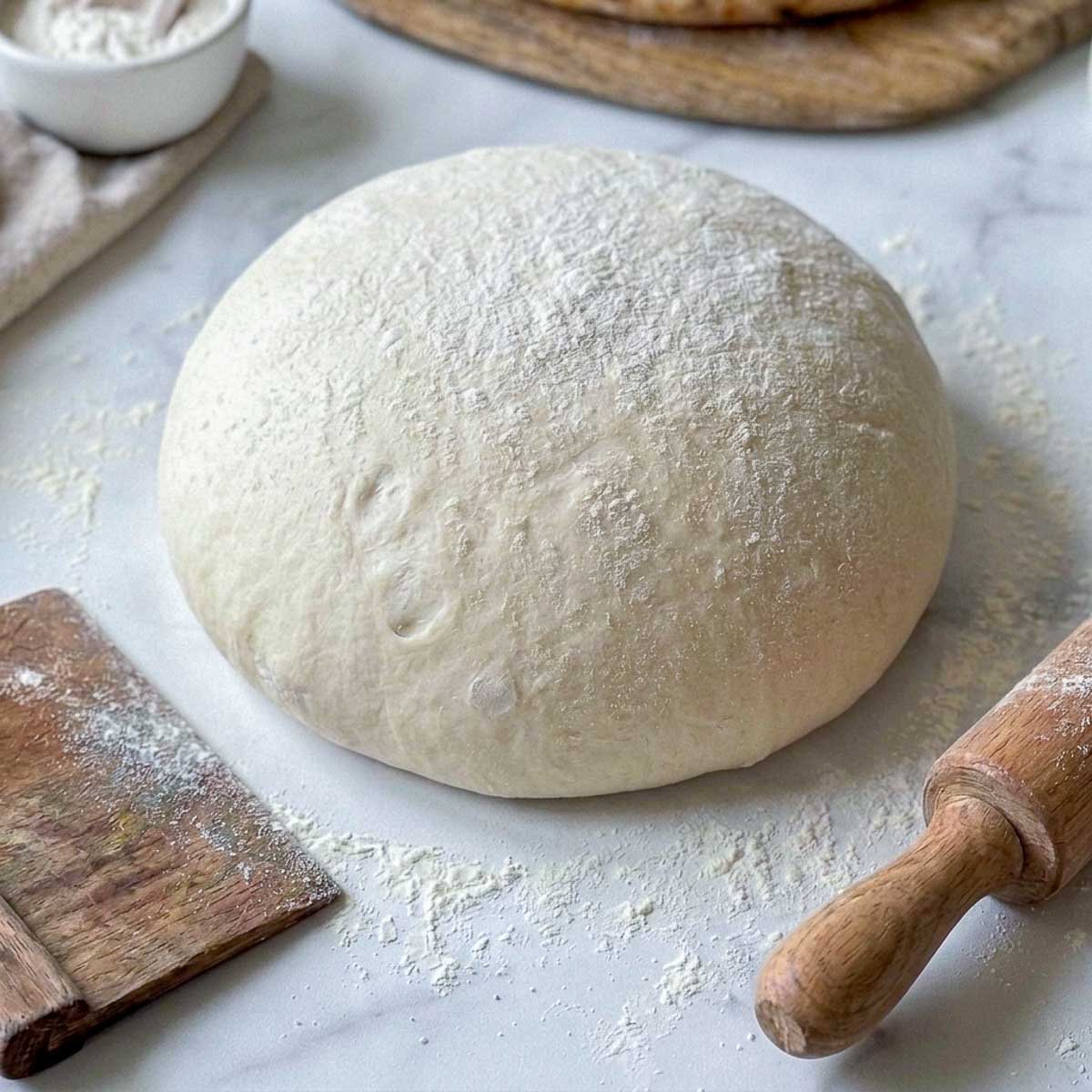 Rested no-yeast kefir pizza dough ball dusted with flour beside a rolling pin and bench scraper.