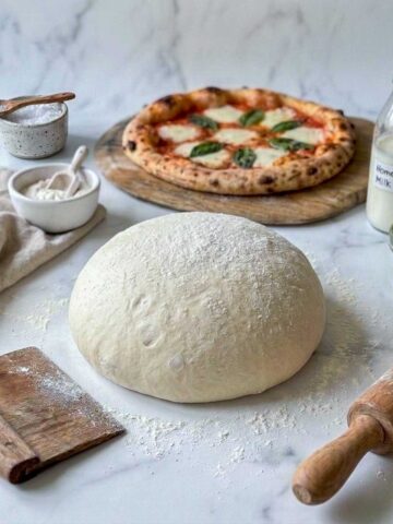 No-yeast kefir pizza dough ball on a floured marble counter with a baked margherita pizza and a bottle of homemade milk kefir.