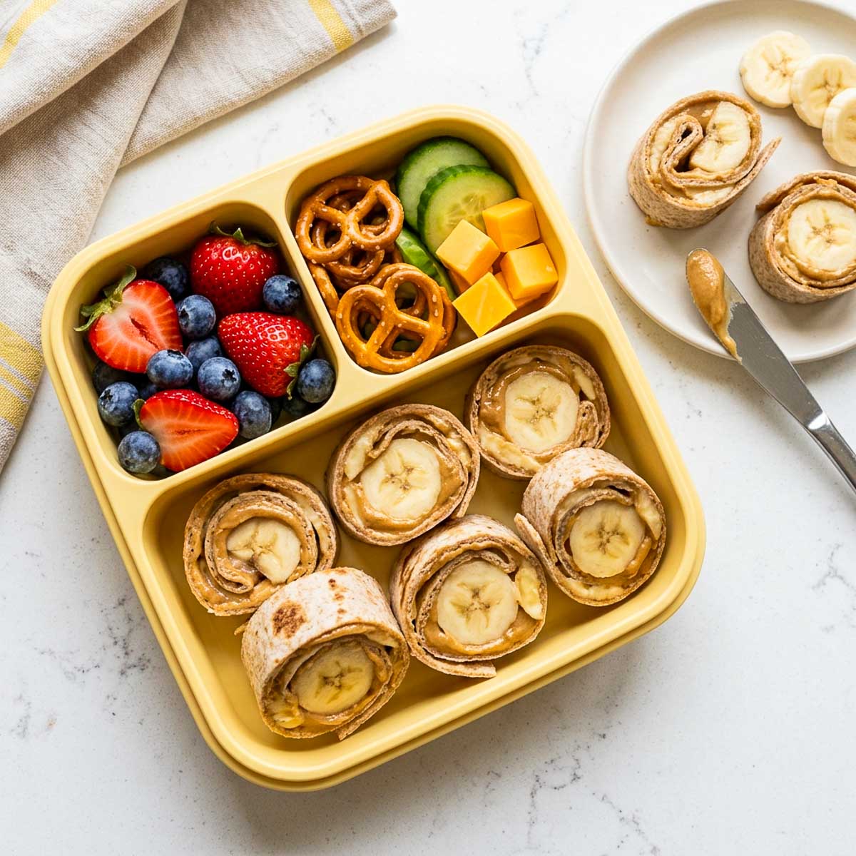 SunButter banana roll-ups packed in a school lunchbox with strawberries, blueberries, pretzels, cucumber, and cheddar cheese.