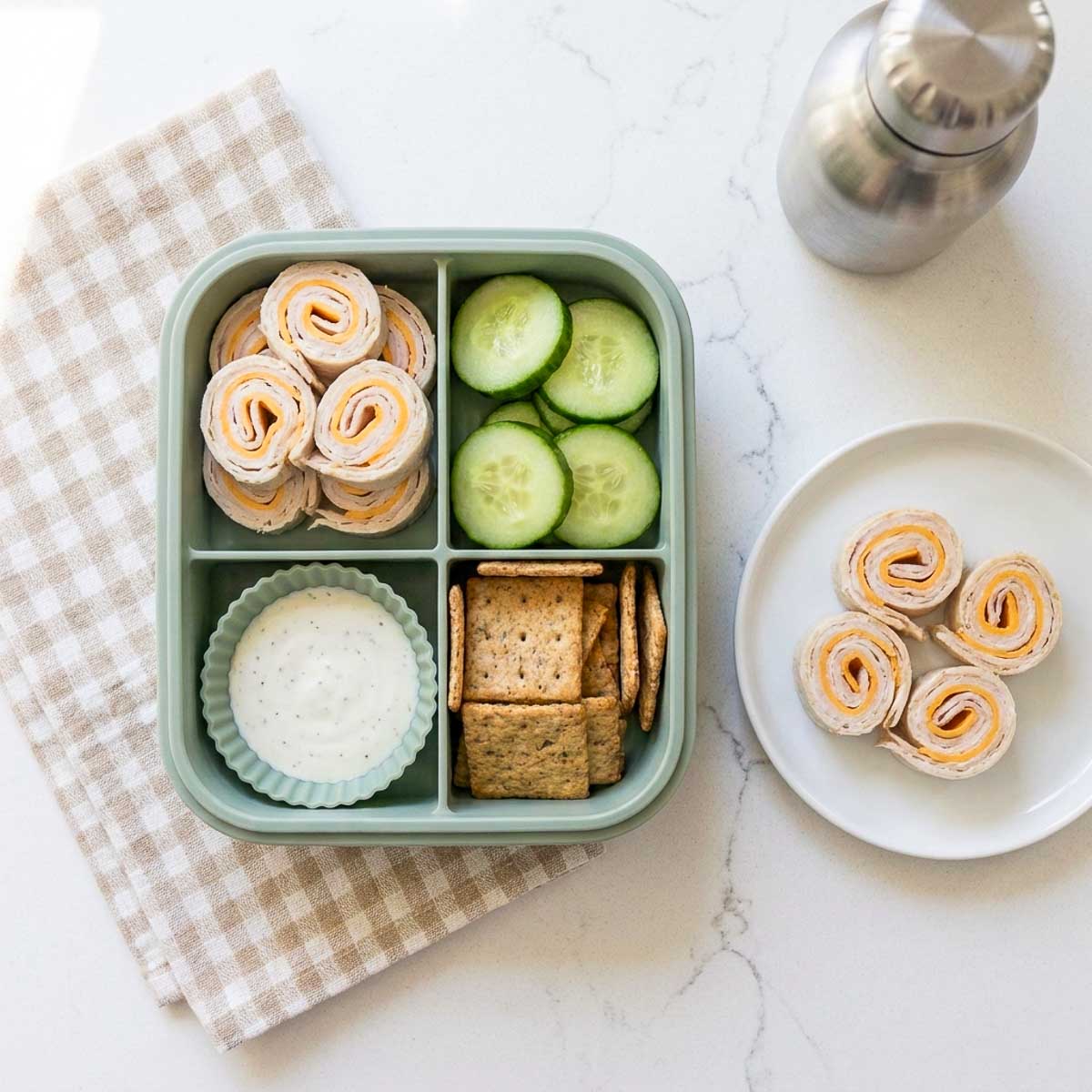Turkey and cheese roll-ups packed in a school lunchbox with cucumber slices, whole-grain crackers, and creamy dip.
