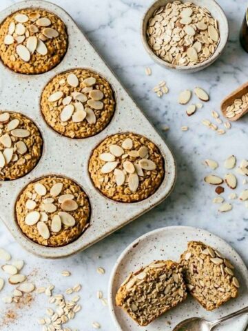 Baked oatmeal cups topped with sliced almonds in a speckled muffin tin, with one cut in half on a plate.