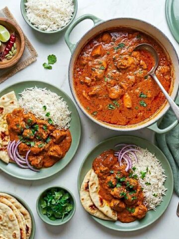 Top-down view of chicken masala served family-style with basmati rice and naan on sage green plates, alongside a Dutch oven of curry, sliced red onions, cilantro, naan, and a sage green linen napkin.